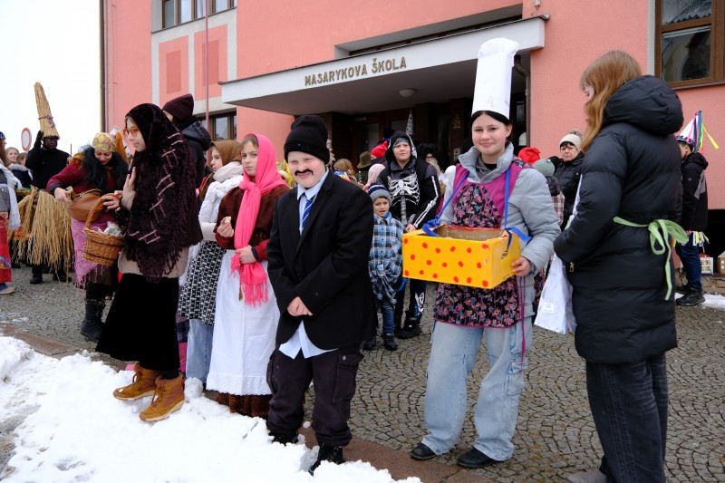 Masopustní masky se shromáždily před školou a čekají na svolení, aby mohly vyrazit k obecnímu úřadu. Mezitím rozdávají výborné sladké koblížky.